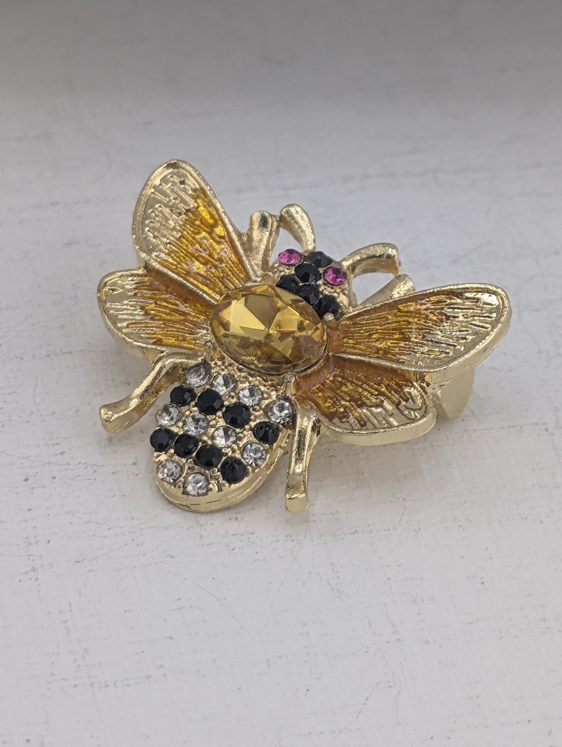 A decorative gold-colored pin or brooch in the shape of a bee, adorned with colorful gemstones and intricate details.