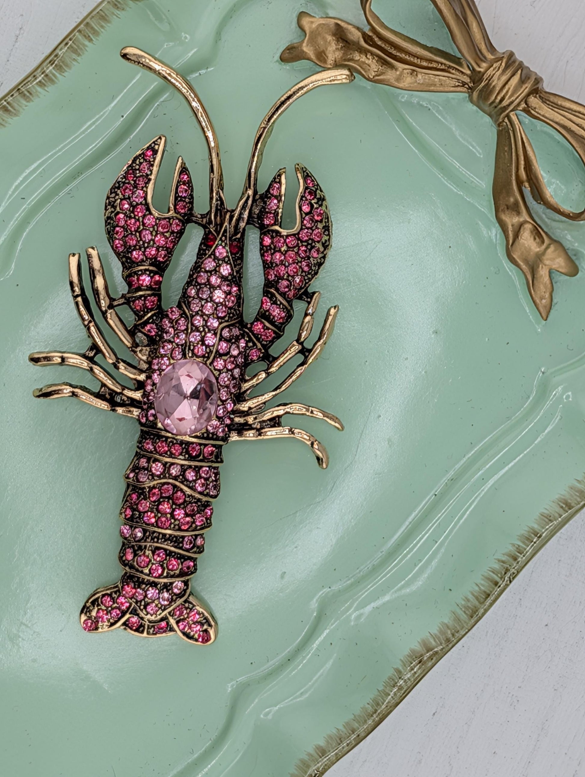 A decorative lobster brooch with pink gemstones and a gold bow is displayed on a green and gold tray.