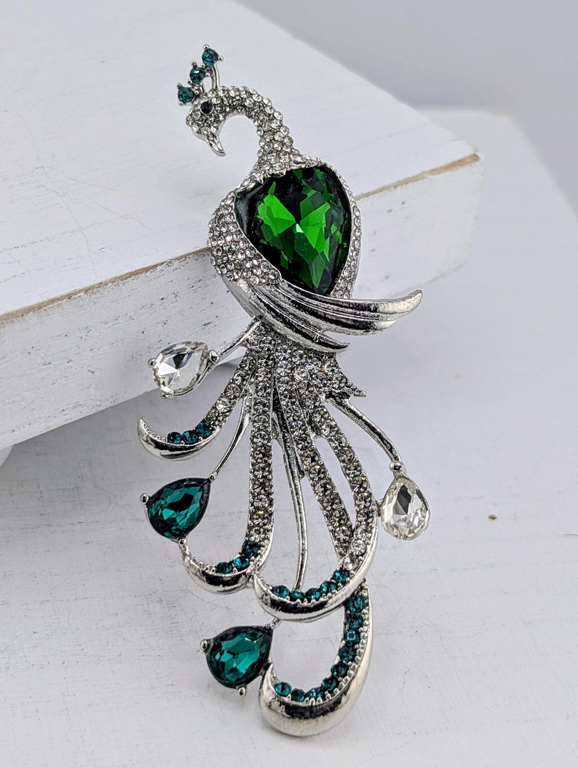 A decorative brooch in the shape of a peacock, adorned with green gemstones and silver rhinestones.