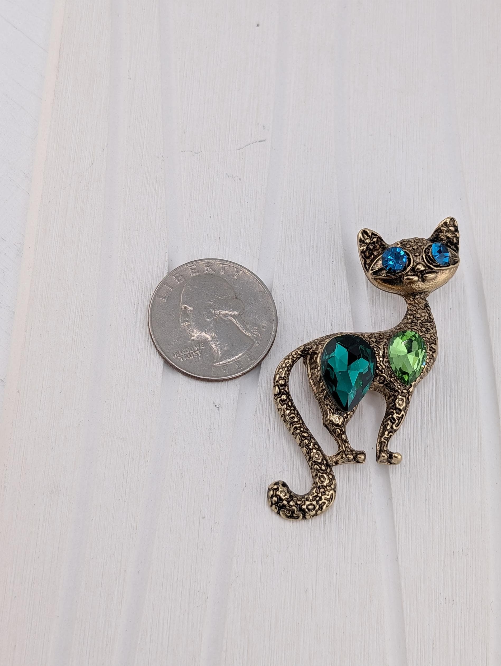 A shiny, ornate cat-shaped brooch made of metal and adorned with colorful gemstones is placed next to a US quarter on a white surface.