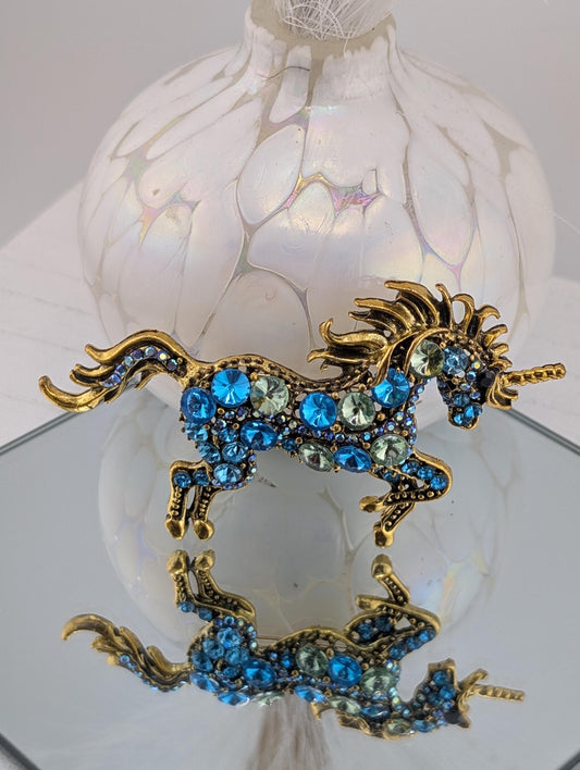 A decorative brooch in the shape of a unicorn, adorned with blue and green gemstones, is displayed on a reflective surface.