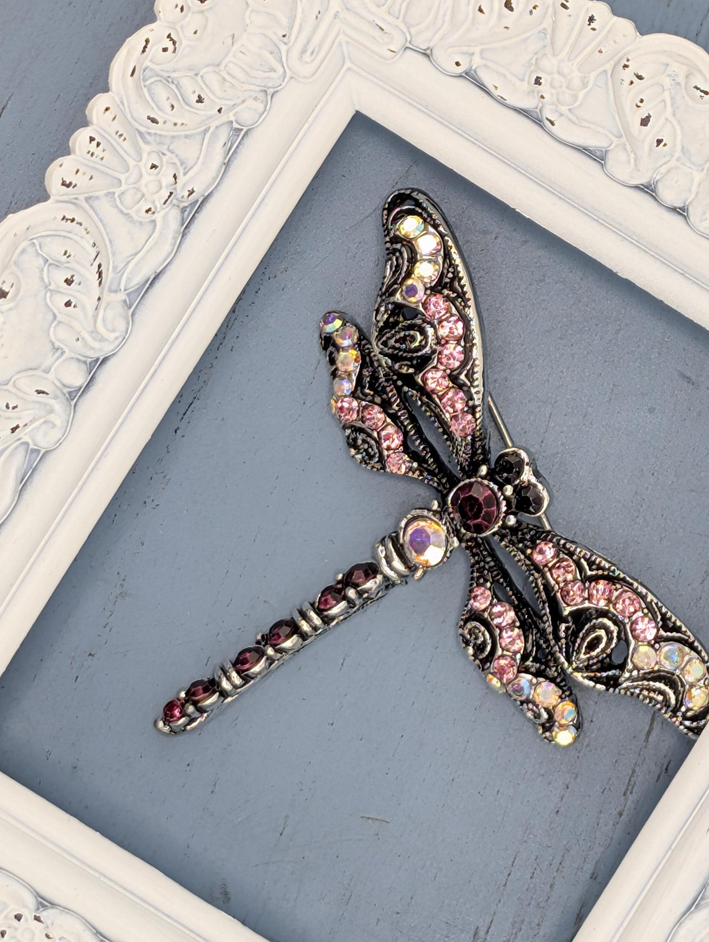 A decorative silver butterfly brooch with pink and purple gemstones is displayed in a white ornate frame against a blue background.