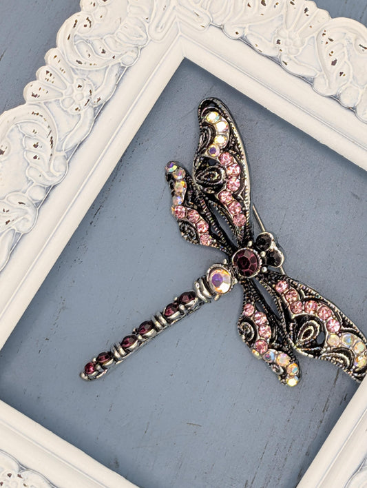 A decorative silver butterfly brooch with pink and purple gemstones is displayed in a white ornate frame against a blue background.