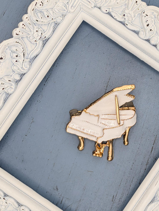 A white and gold piano brooch is displayed on a blue surface, framed by a white ornate frame.