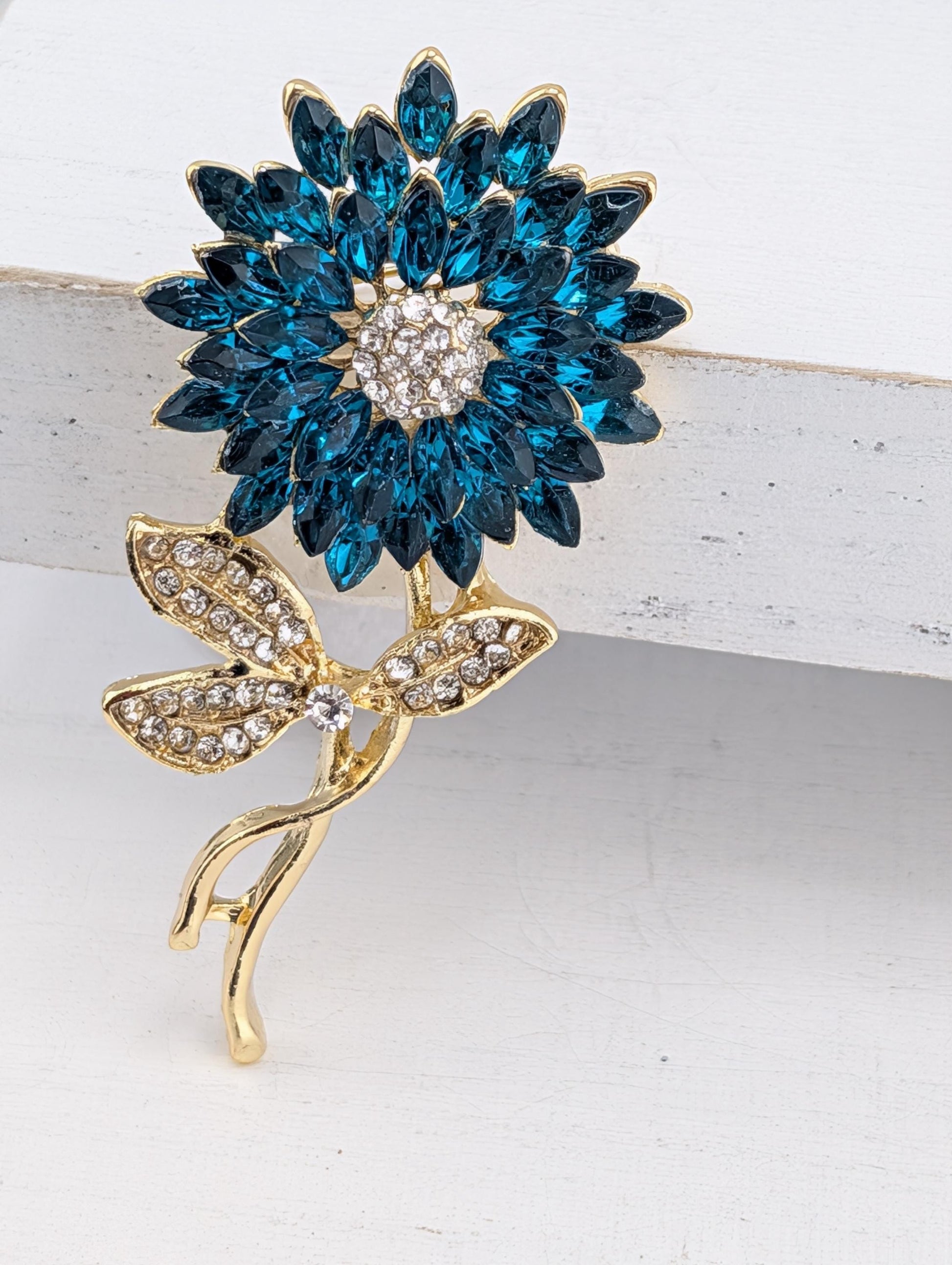 A decorative brooch in the shape of a blue flower with gold leaves and a large central gemstone.