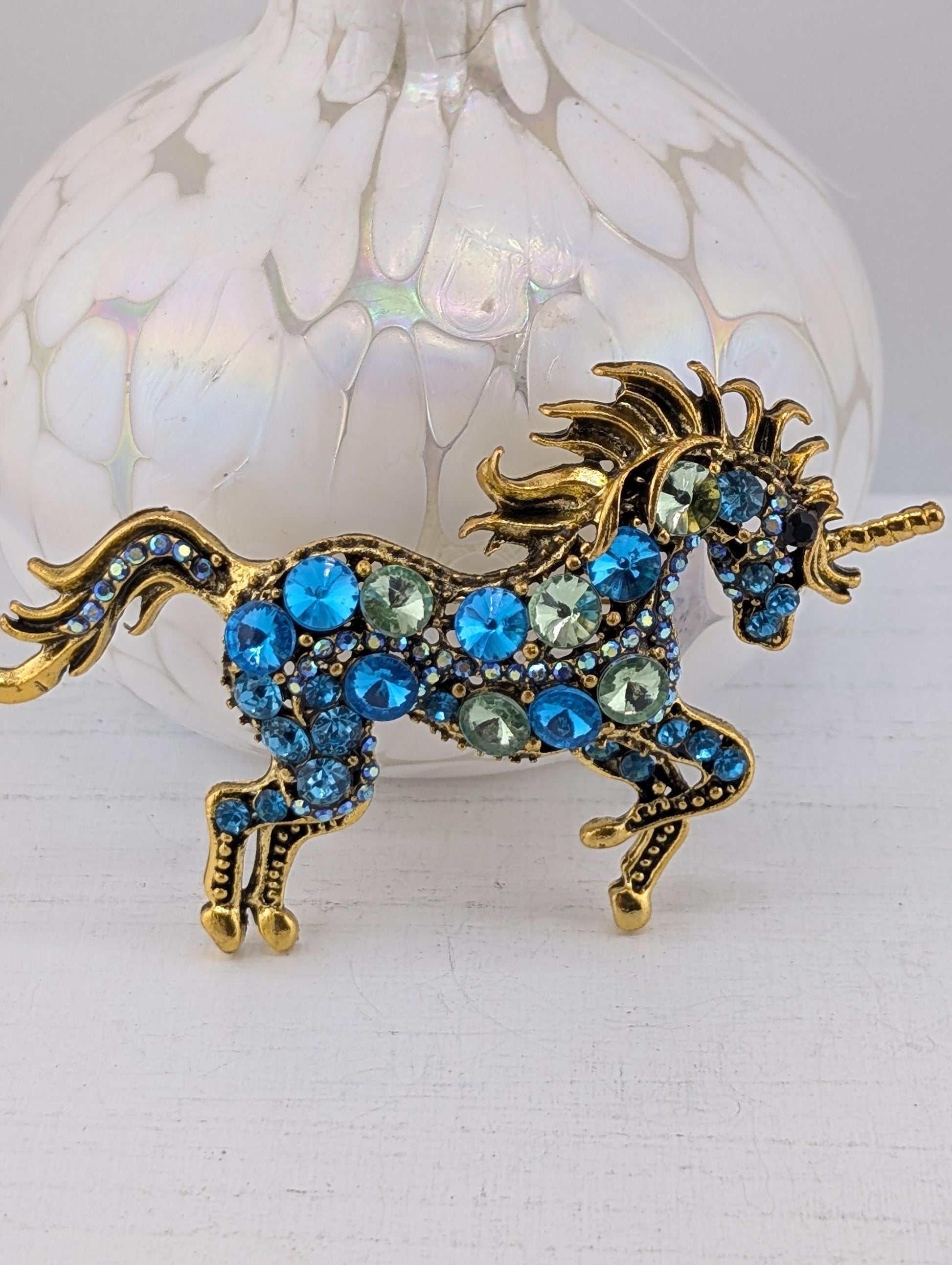 A decorative brooch in the shape of a unicorn, adorned with blue and green gemstones, is displayed against a white background.
