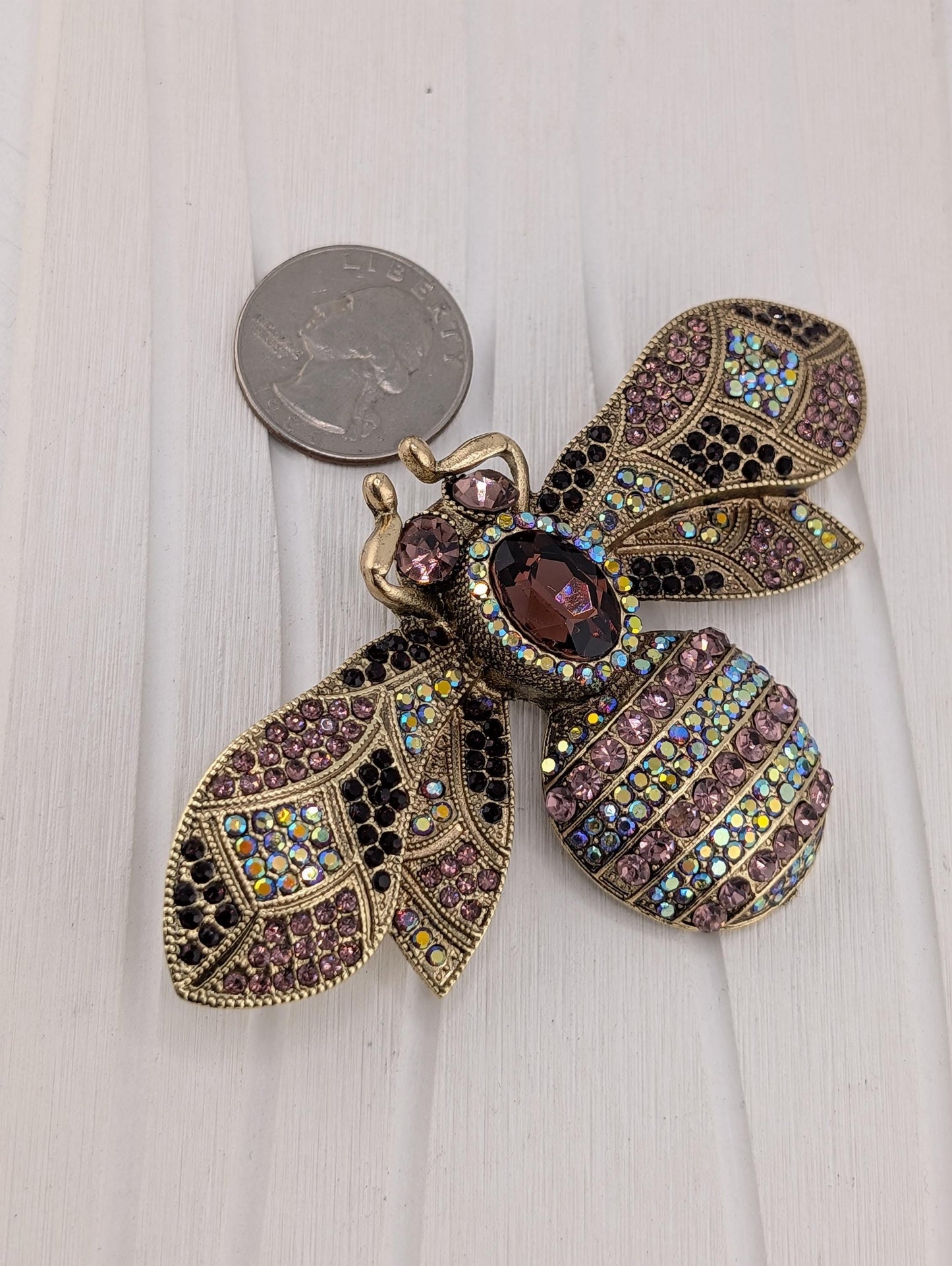 A decorative brooch in the shape of a bee, adorned with colorful gemstones and intricate details, is placed on a white surface next to a coin.