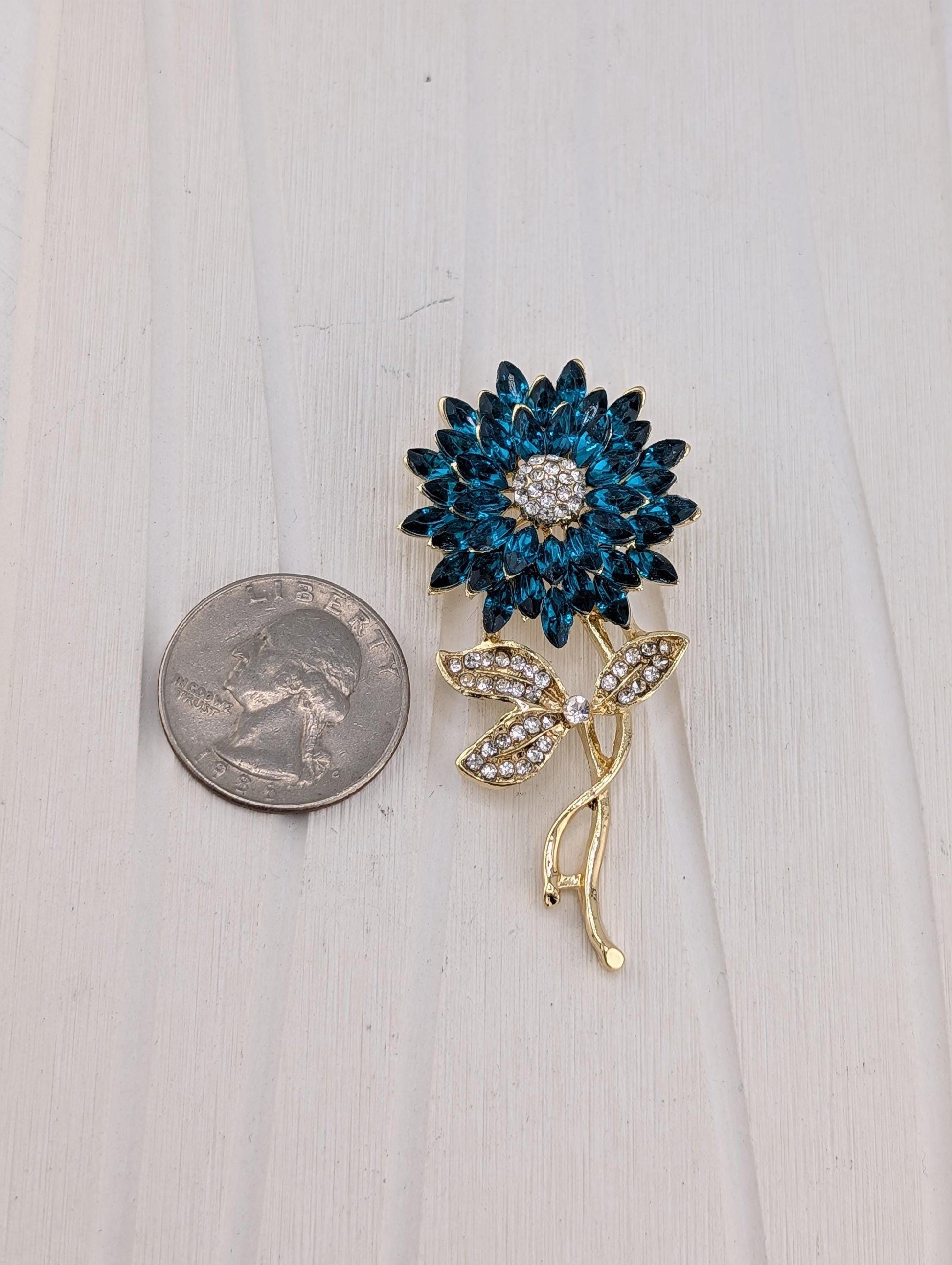 A blue flower brooch with a gold stem and leaves, adorned with sparkling gemstones, is placed next to a United States quarter on a white surface.