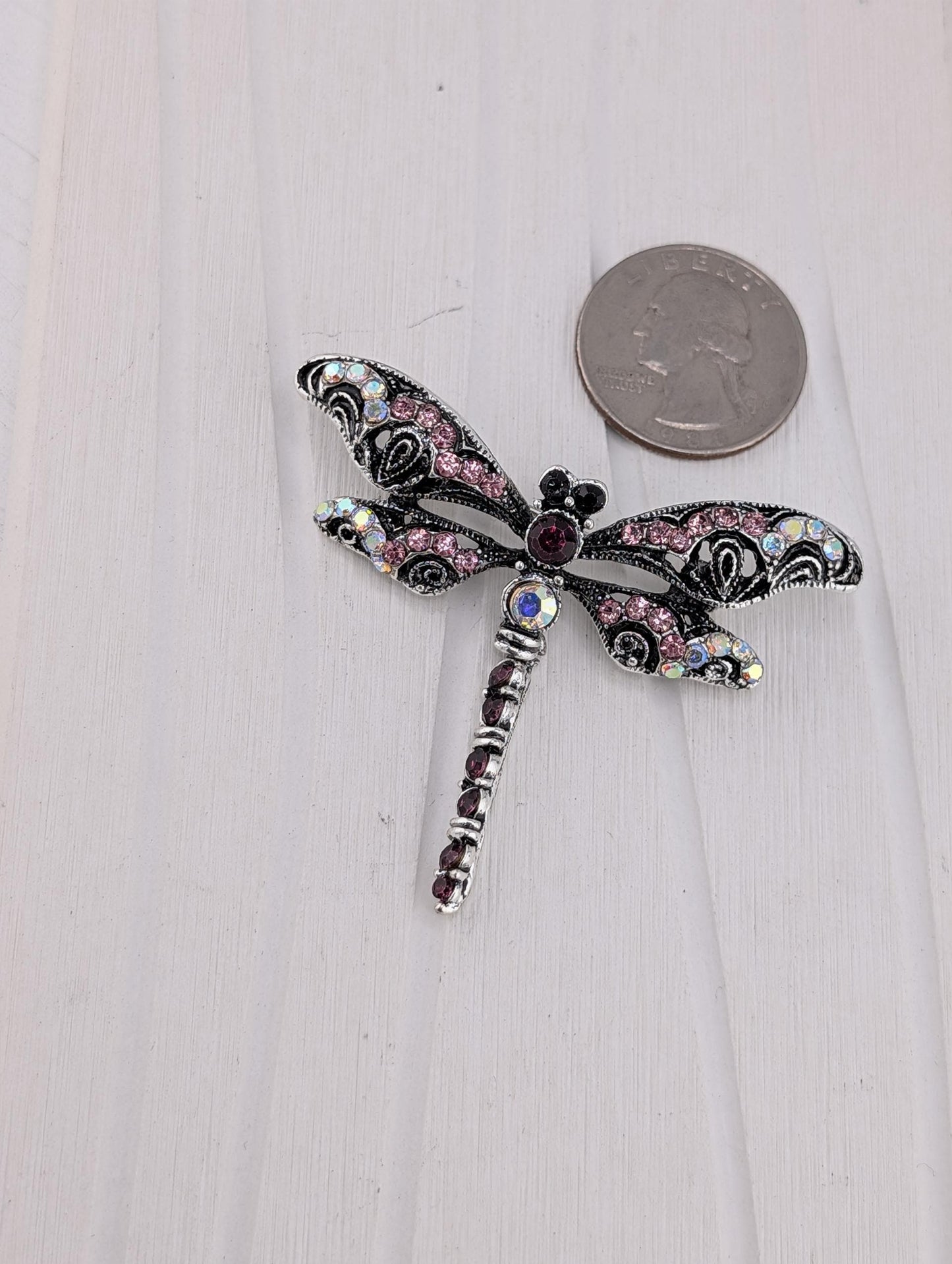 A decorative silver-toned brooch in the shape of a dragonfly, adorned with colorful gemstones and a central eye-shaped gemstone. The brooch is placed on a white wooden surface next to a coin.