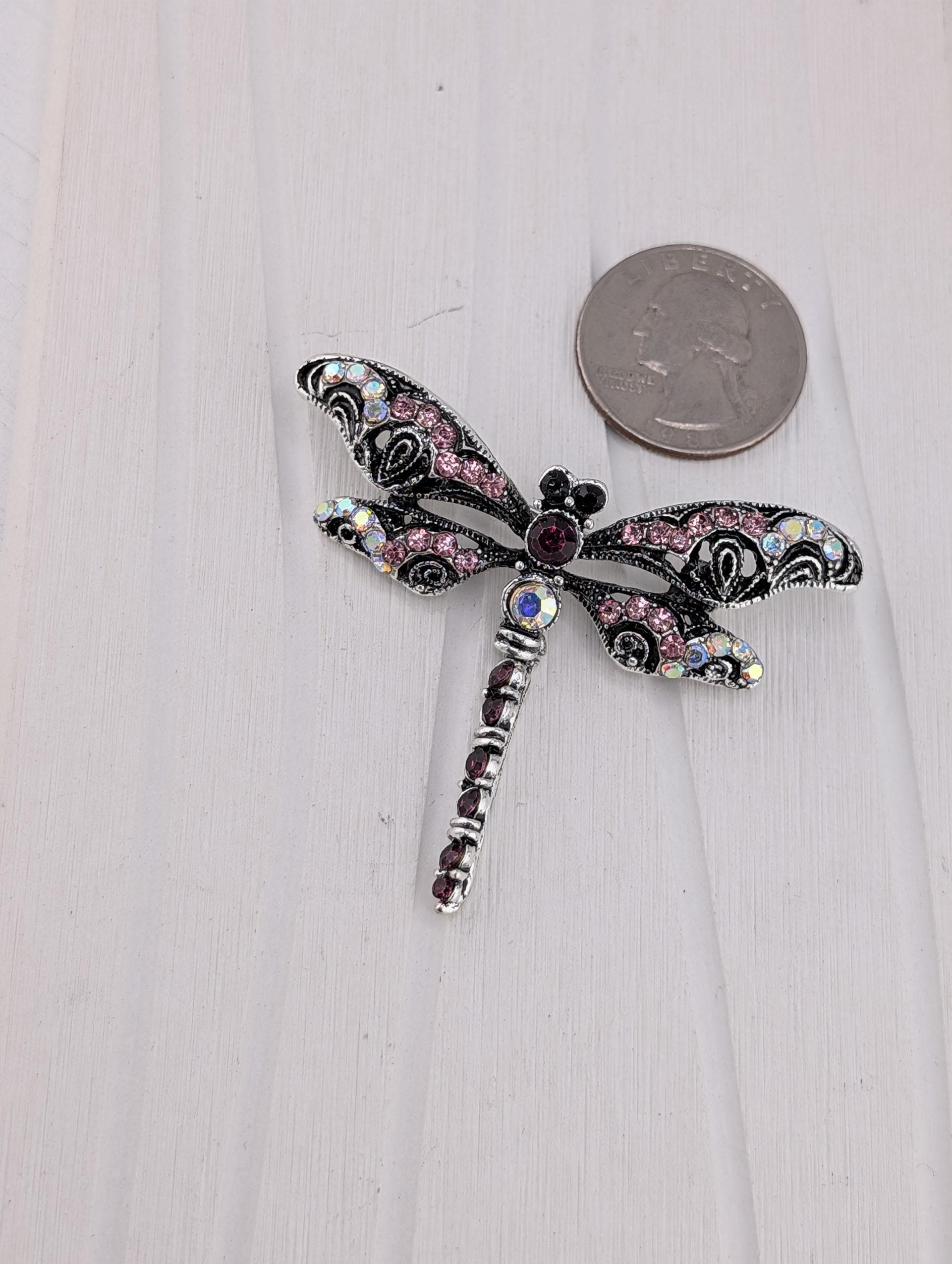 A decorative silver-toned brooch in the shape of a dragonfly, adorned with colorful gemstones and a central eye-shaped gemstone. The brooch is placed on a white wooden surface next to a coin.