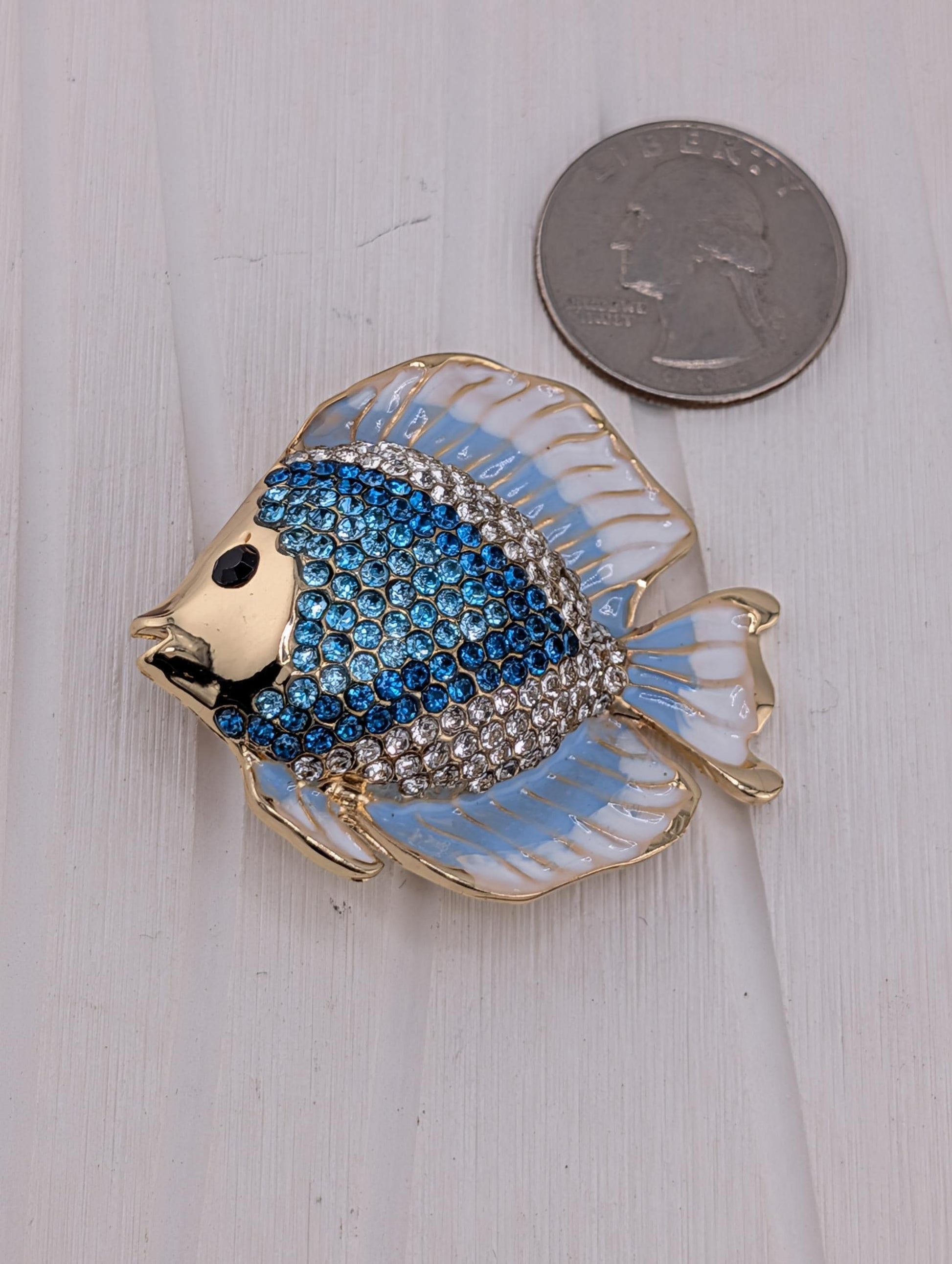 A decorative brooch in the shape of a blue and white fish, adorned with rhinestones, is placed next to a United States quarter coin.