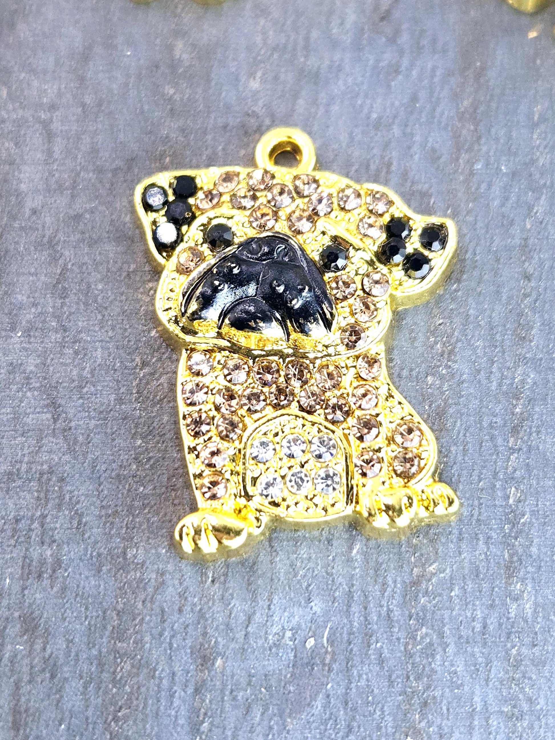 A gold-colored charm in the shape of a dog, adorned with small diamonds and rhinestones, is displayed on a gray surface.