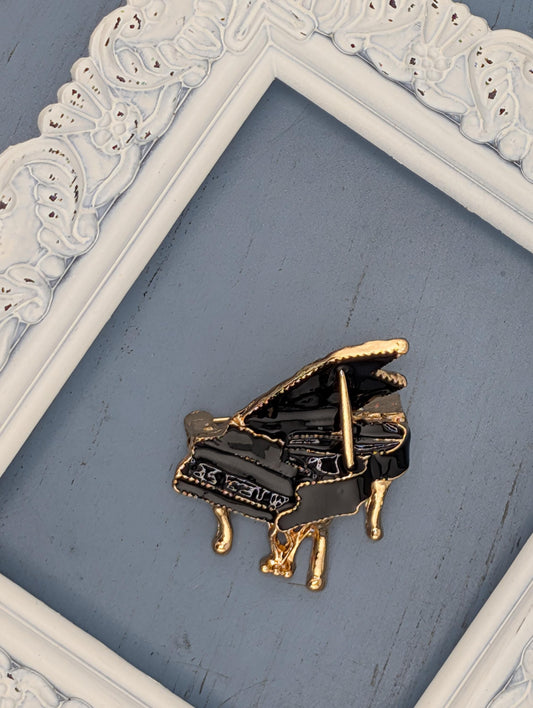 A decorative gold-colored piano brooch is displayed on a blue surface, framed by a white ornate frame.