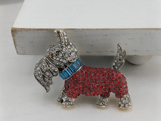 A decorative dog brooch with a red body and a blue collar, adorned with sparkling rhinestones.