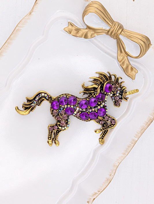A decorative piece featuring a purple unicorn adorned with numerous purple gemstones, placed on a white surface with a gold bow.
