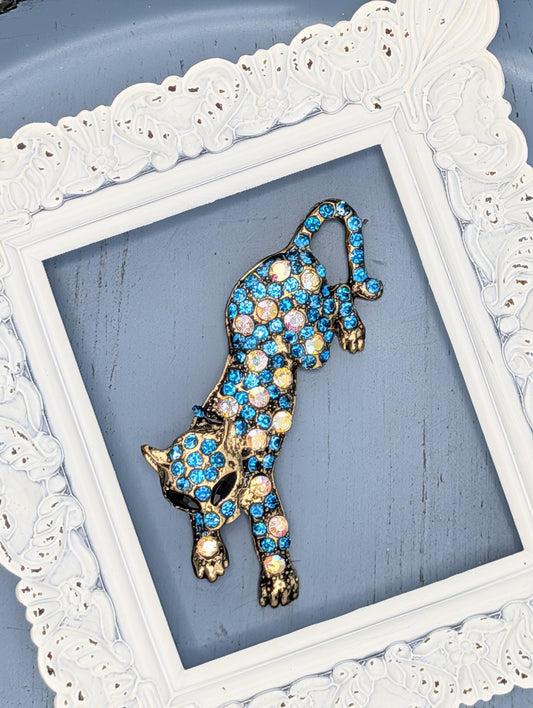 A decorative cat brooch with blue and gold gemstones is displayed in a white ornate frame.