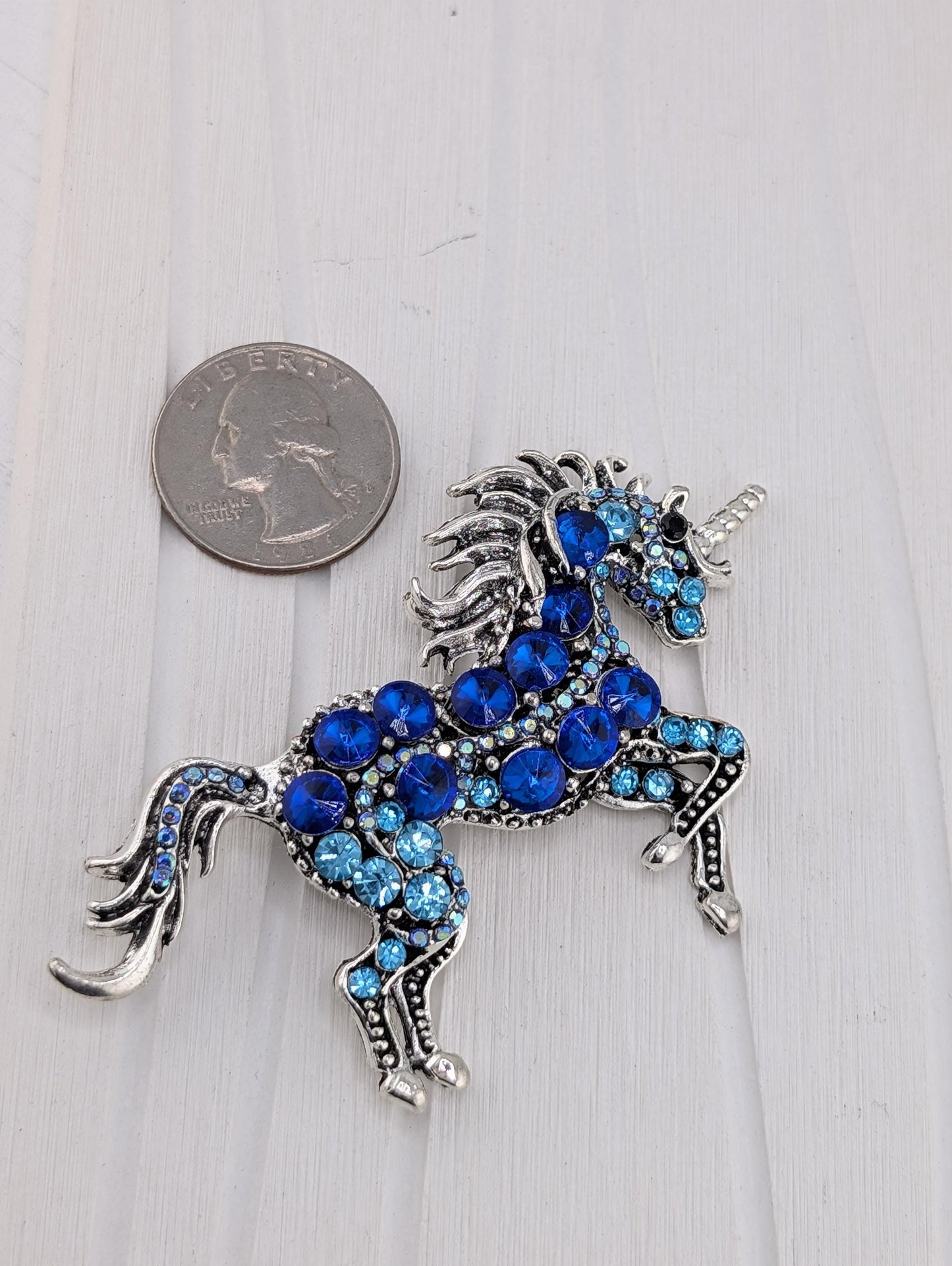 A silver-colored brooch in the shape of a unicorn, adorned with blue gemstones, is placed next to a United States quarter coin on a white surface.