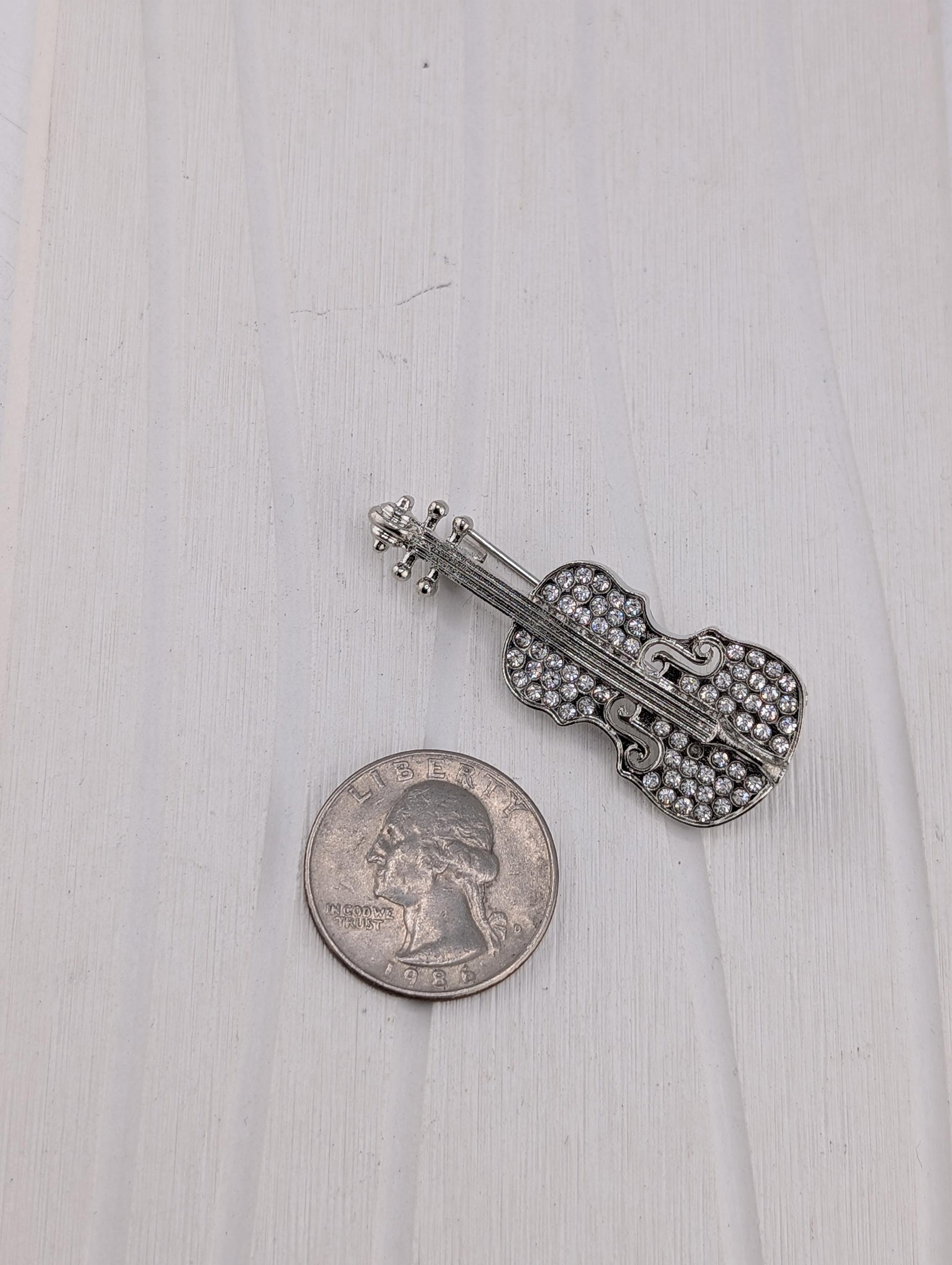 A silver-colored violin brooch with rhinestones is placed next to a United States quarter on a white wooden surface.