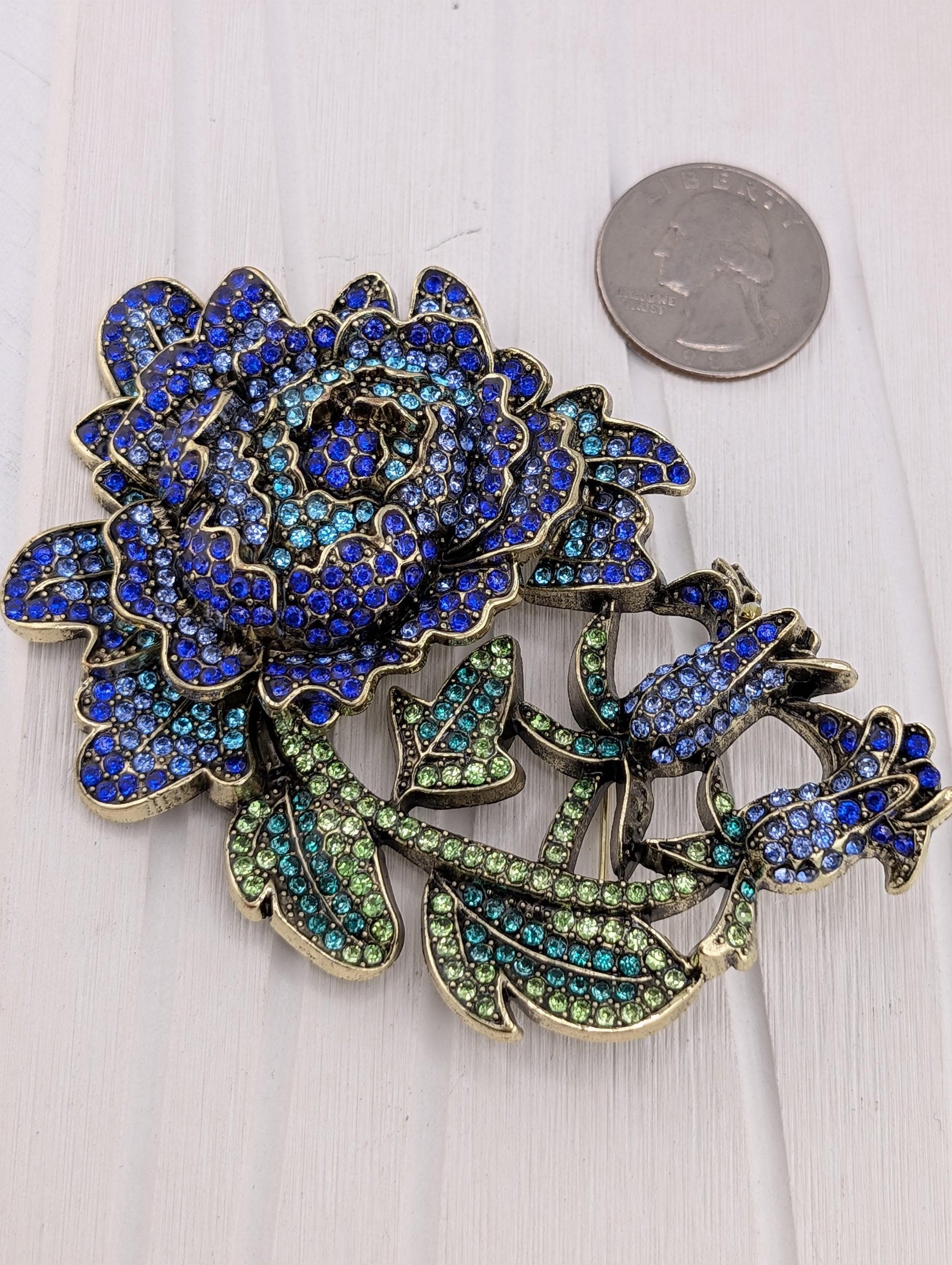 A decorative brooch with a large blue flower and green leaves, adorned with sparkling rhinestones.