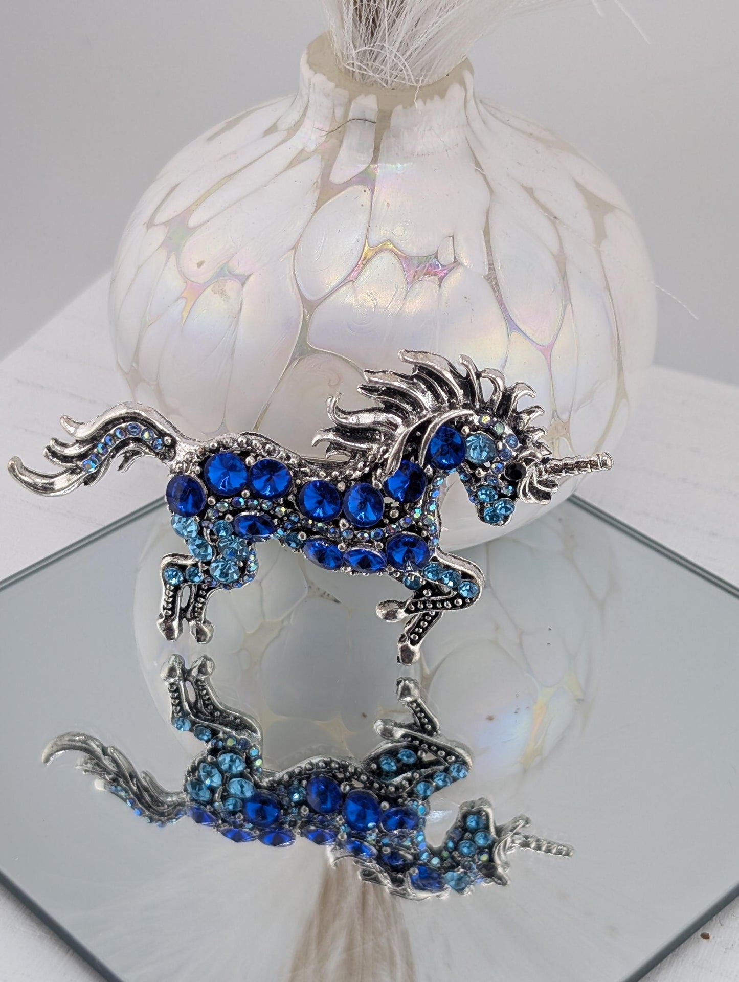 A decorative blue unicorn brooch with blue gemstones is displayed on a white surface, with a white vase and a mirror reflecting the unicorn and the vase.