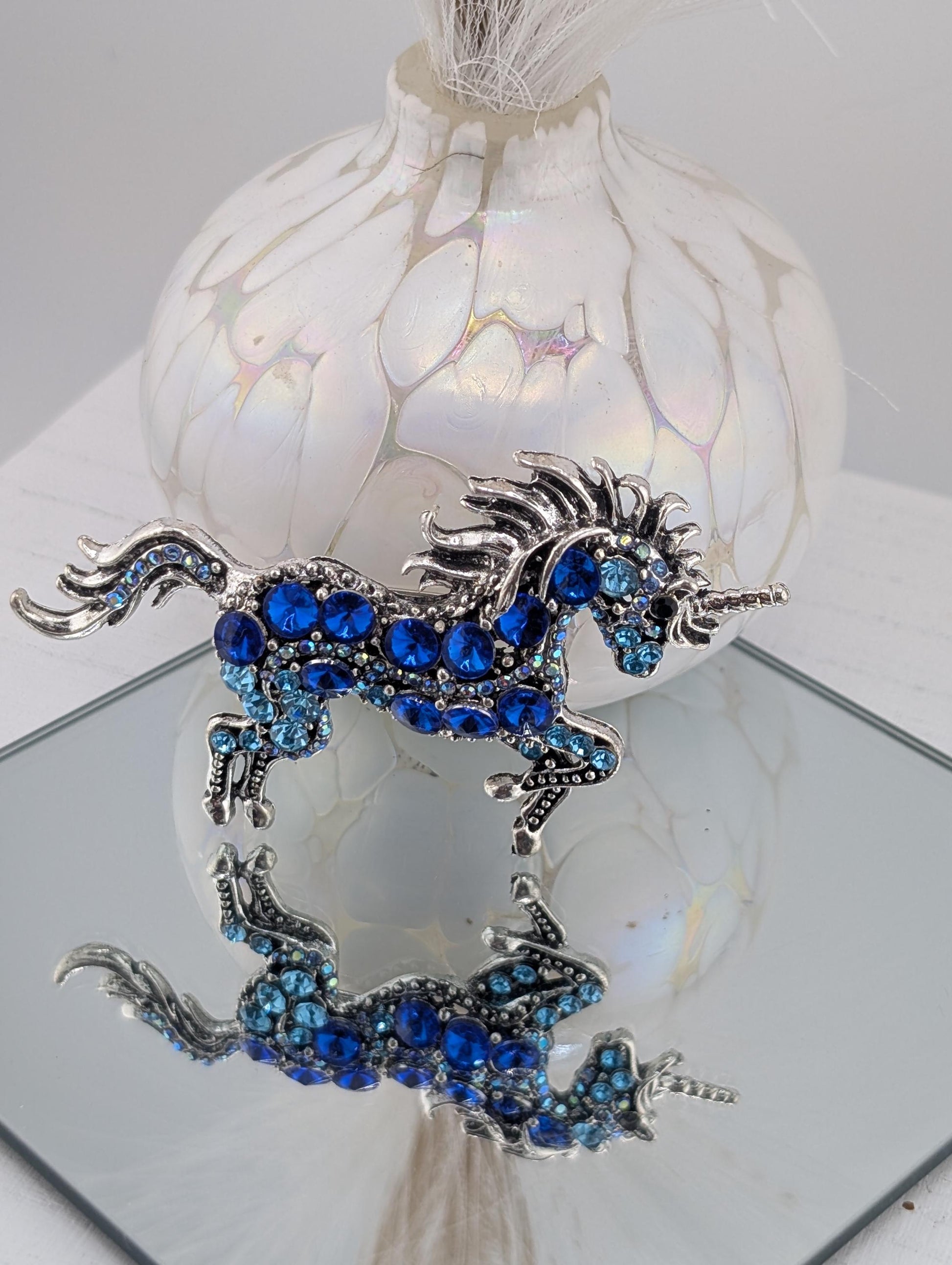 A decorative blue unicorn brooch with blue gemstones is displayed on a white surface, with a white vase and a mirror reflecting the unicorn and the vase.