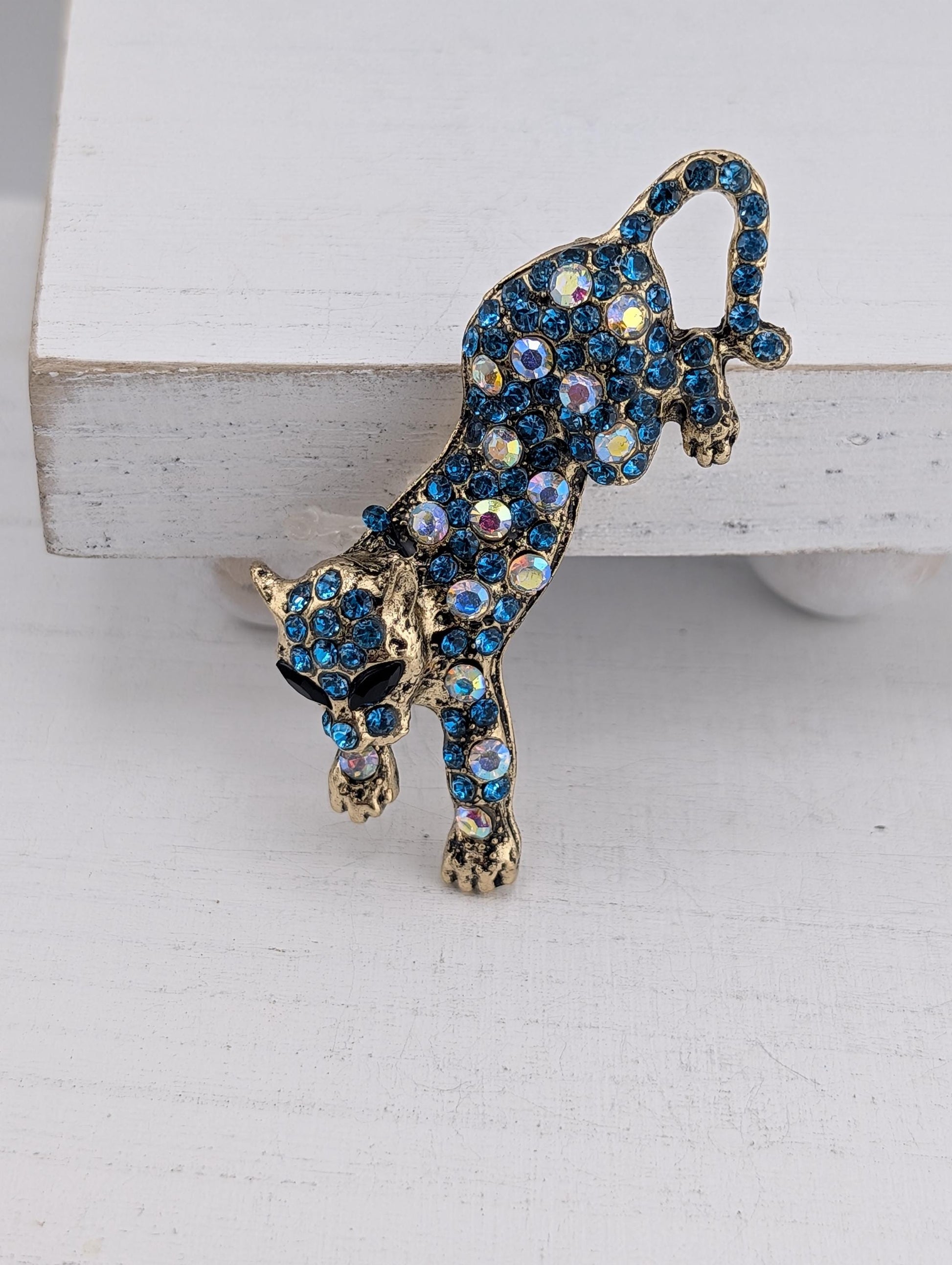 A decorative brooch in the shape of a cat, adorned with blue and gold gemstones, is displayed on a white surface.
